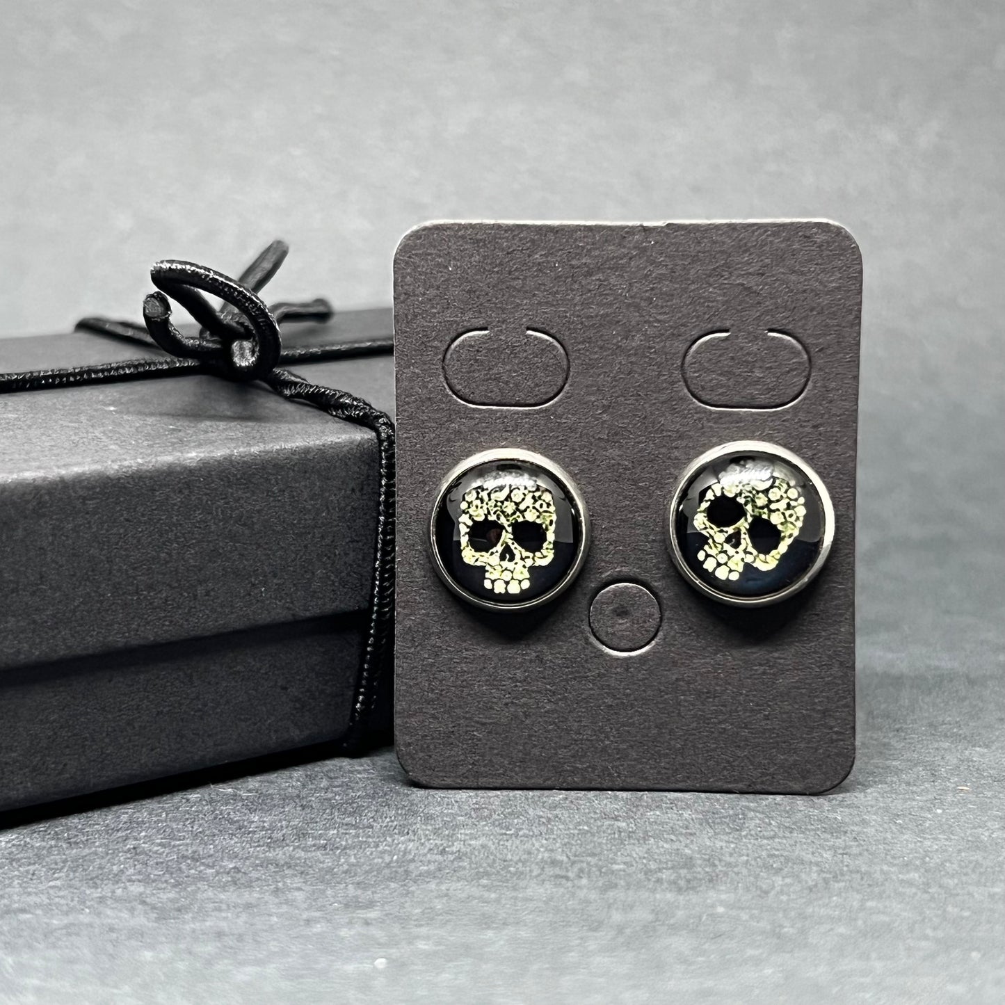 Men's skull stud earrings with dark black and bone color tone designs on silver frame with clear acrylic glass. Man cacochon earrings. Rock, Punk, and Tattoo fashion for man and guys by CALACAMANIA™ accessories.