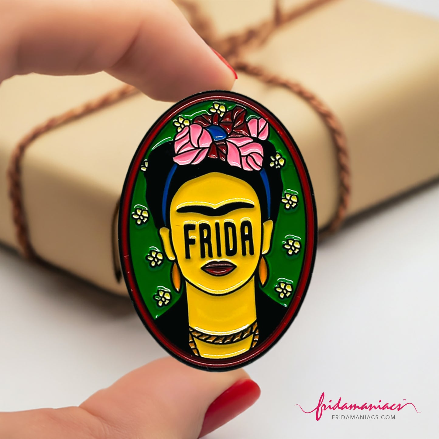 Frida Kahlo oval enamel pin back button with green enamel as background, wine tone frame, and FRIDA spelling instead of eyes below iconic artist eyebrows with pink flowers. Fridamaniacs. Mexican jewelry.