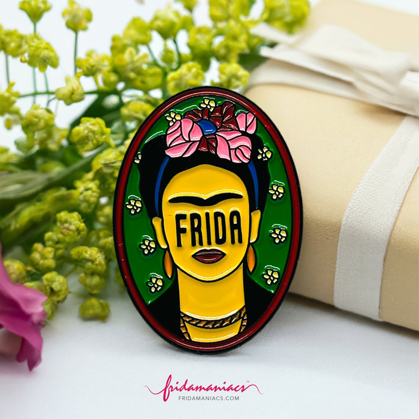 Frida Kahlo oval enamel pin back button with green enamel as background, wine tone frame, and FRIDA spelling instead of eyes below iconic artist eyebrows with pink flowers. Fridamaniacs. Mexican jewelry.