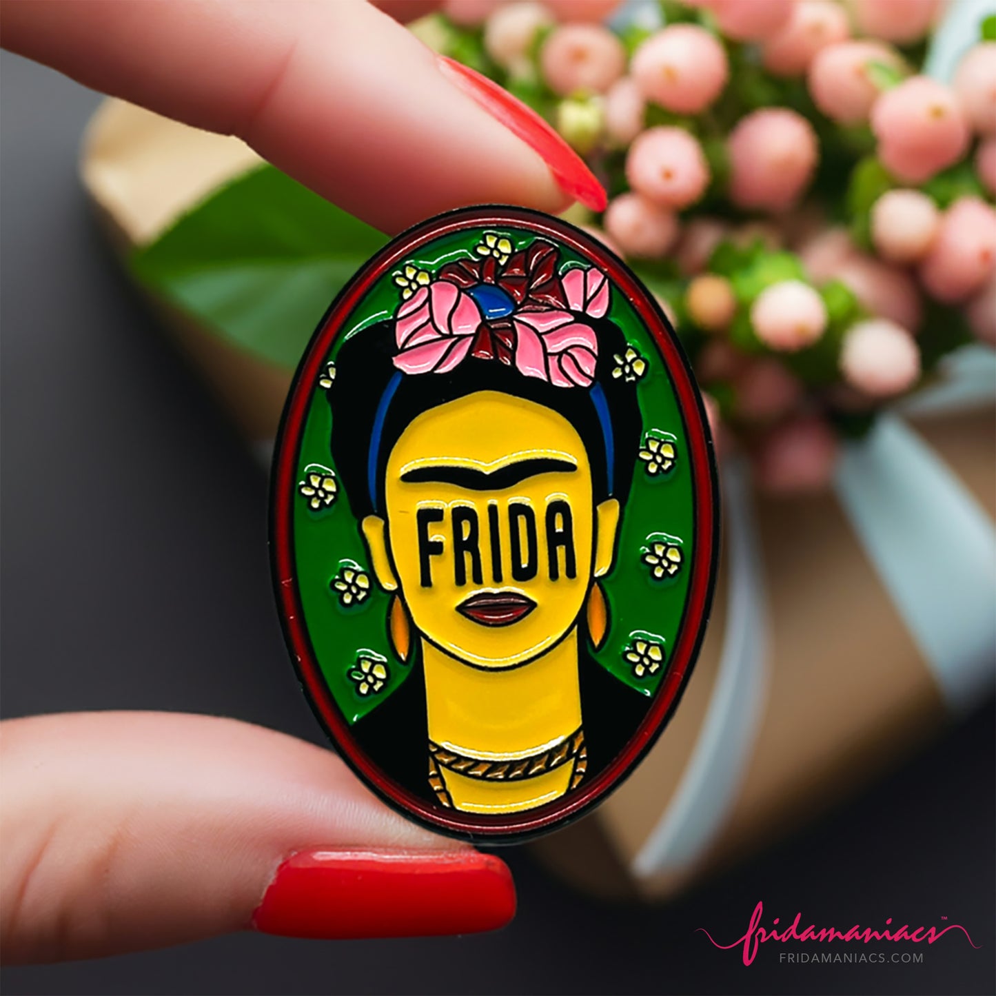 Frida Kahlo oval enamel pin back button with green enamel as background, wine tone frame, and FRIDA spelling instead of eyes below iconic artist eyebrows with pink flowers. Fridamaniacs. Mexican jewelry.