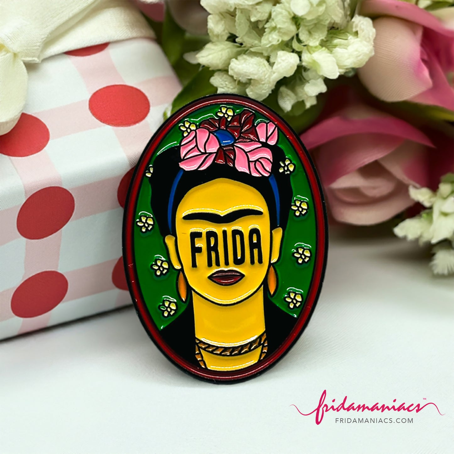 Frida Kahlo oval enamel pin back button with green enamel as background, wine tone frame, and FRIDA spelling instead of eyes below iconic artist eyebrows with pink flowers. Fridamaniacs. Mexican jewelry.