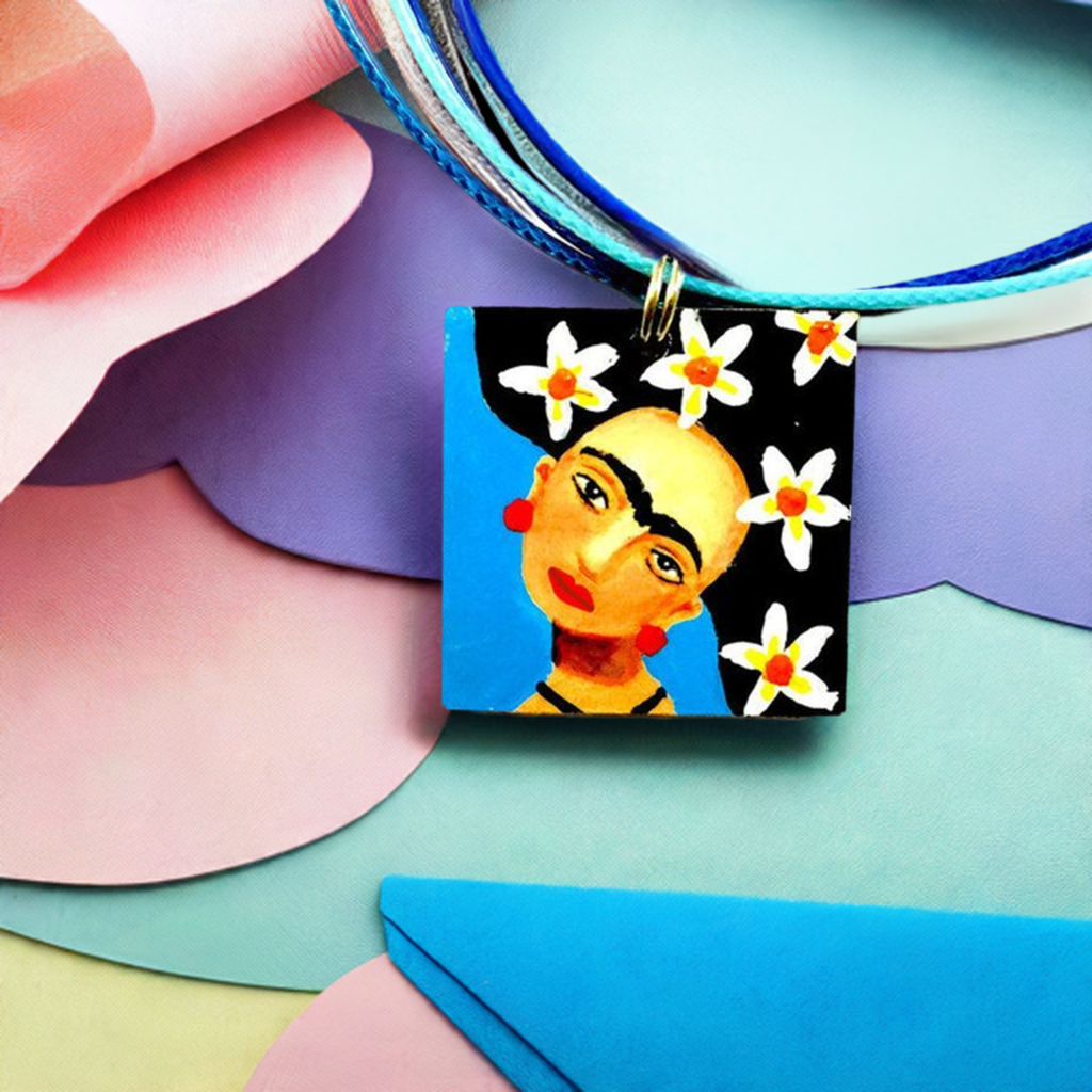 Hand painted Frida Kahlo inspired pendant with blue and white-yellow-orange wilds flowers on hair. Mexican artist portrait art to wear for women and girls. Unique gift idea for fridamaniacs, fridalovers, and frida fans.
