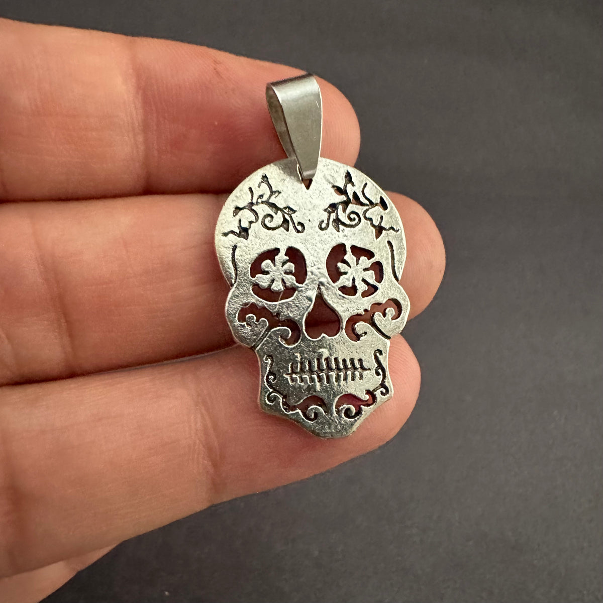 Close-up of the skull pendant held against fingers, showing its silver-tone bail, textured surface, floral eyes, and curling cheek details.
