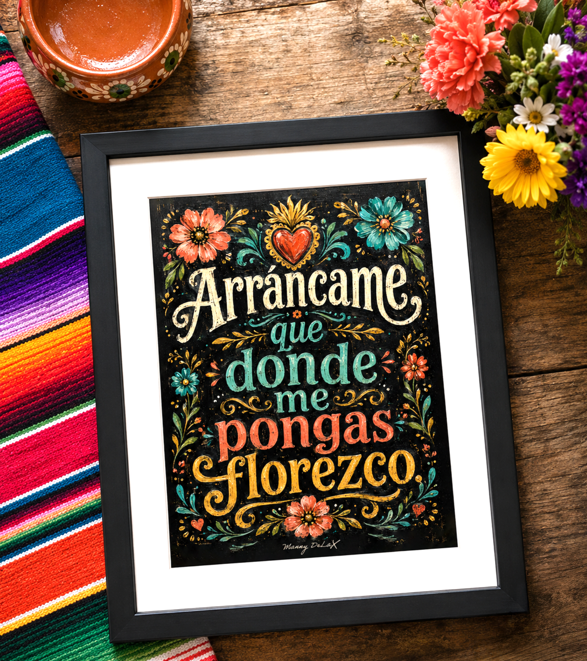 A framed art print featuring the Spanish phrase 'Arráncame que donde me pongas florezco' in decorative multicolor lettering on a black background with floral and ornamental illustrations. The print is shown in a black frame with a white mat, placed on a rustic wooden surface next to a colorful striped textile and flowers.