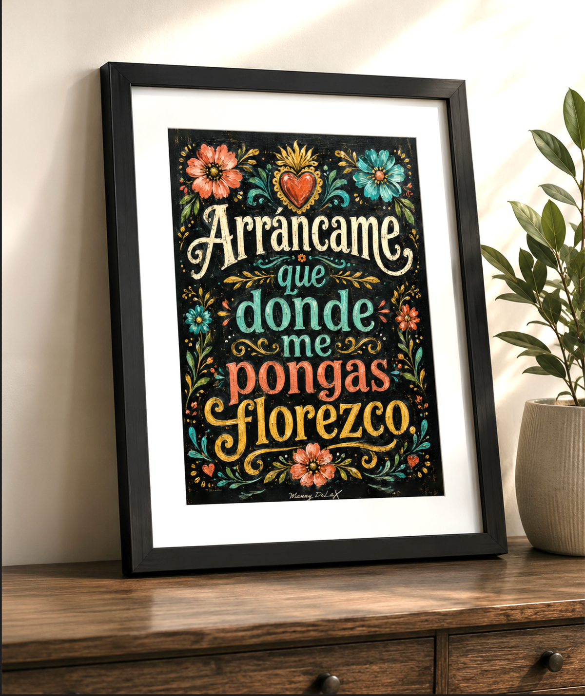 A framed decorative wall art print with Spanish text and colorful floral illustrations, leaning on a wooden surface against a light wall next to a potted plant.