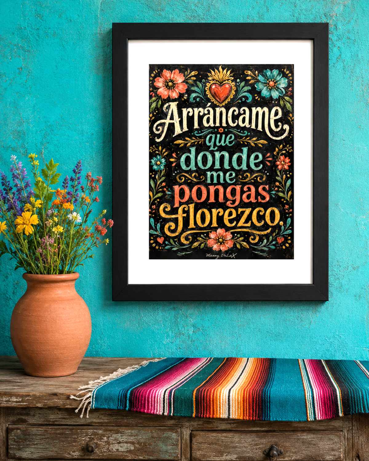 Framed wall art featuring the Spanish phrase 'Arrancame que donde me pongas floresco' in decorative multicolor lettering on a black background with floral illustrations. The frame hangs on a turquoise wall above a wooden surface with a terracotta vase of flowers and a striped serape blanket.