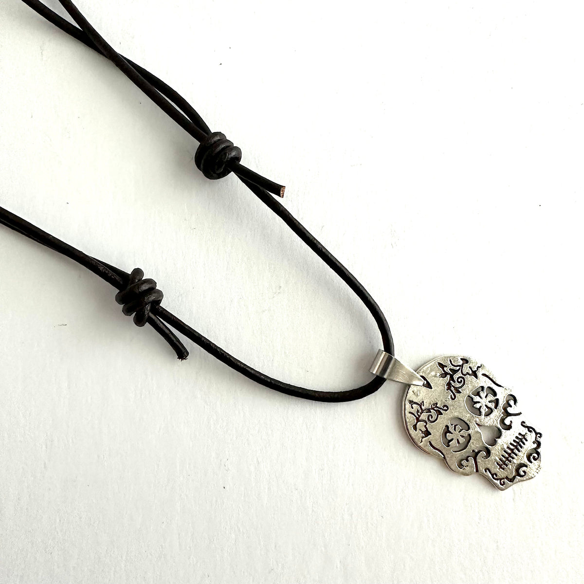 Angled close-up of the sugar skull necklace on white, showing both leather adjustment knots and the decorative silver-tone pendant.