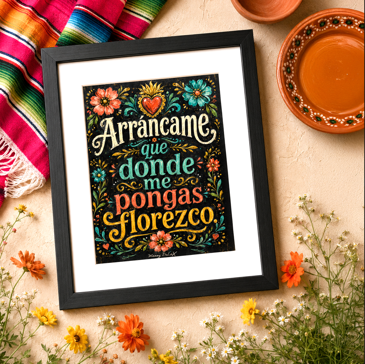 A framed art print featuring the Mexican folk song 'Arráncame que donde me pongas flores' in a black frame, styled on a beige surface with a colorful serape blanket, terracotta dishes, and small yellow and orange flowers.