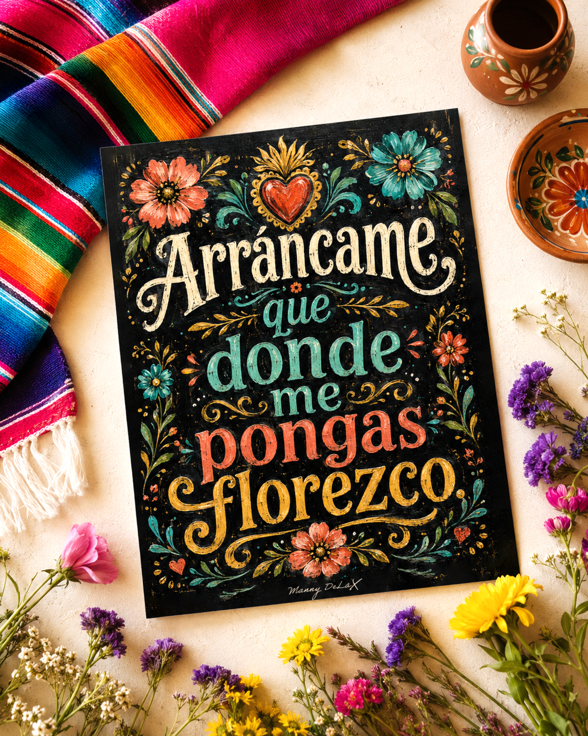 A decorative art print with the Spanish phrase 'Arrancame que donde me pongas florezco' in stylized lettering, surrounded by colorful floral illustrations on a black background. The print is styled in a flat-lay arrangement with a striped serape blanket, small clay pots, and dried flowers on a light beige surface.