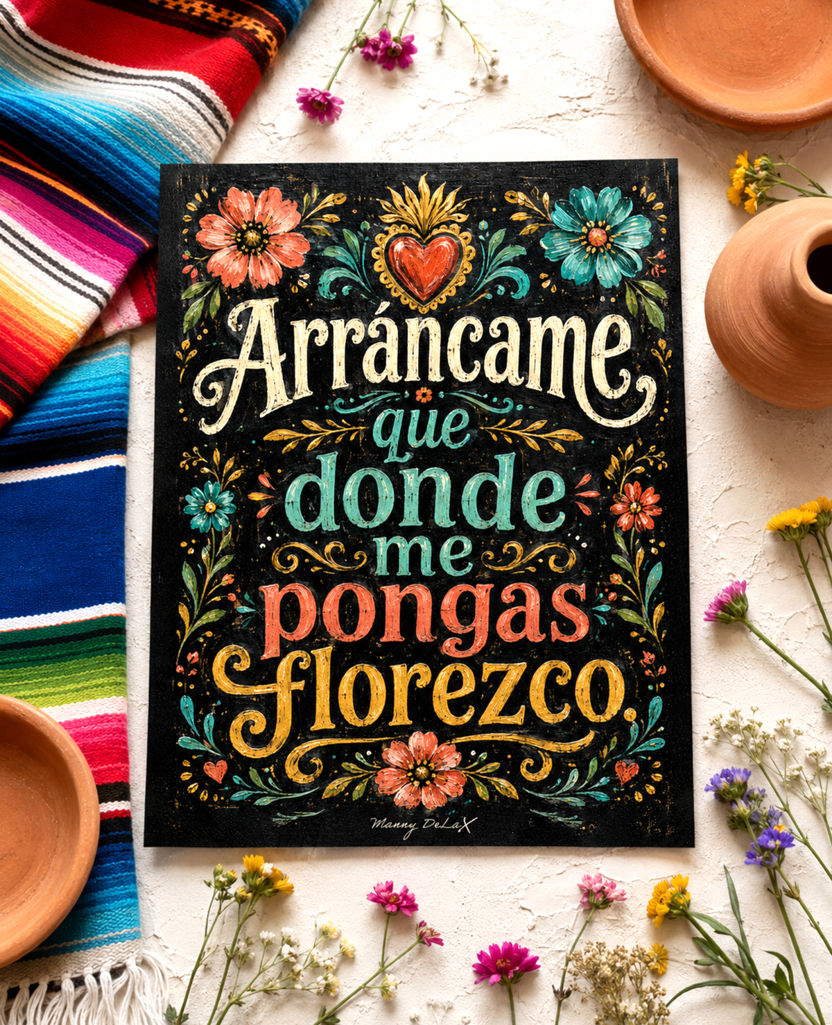 A rectangular black art print with colorful hand-lettered Spanish text reading 'Arráncame que donde me pongas flores' surrounded by floral illustrations, styled on a light surface with a striped textile, bowls, and small flowers.