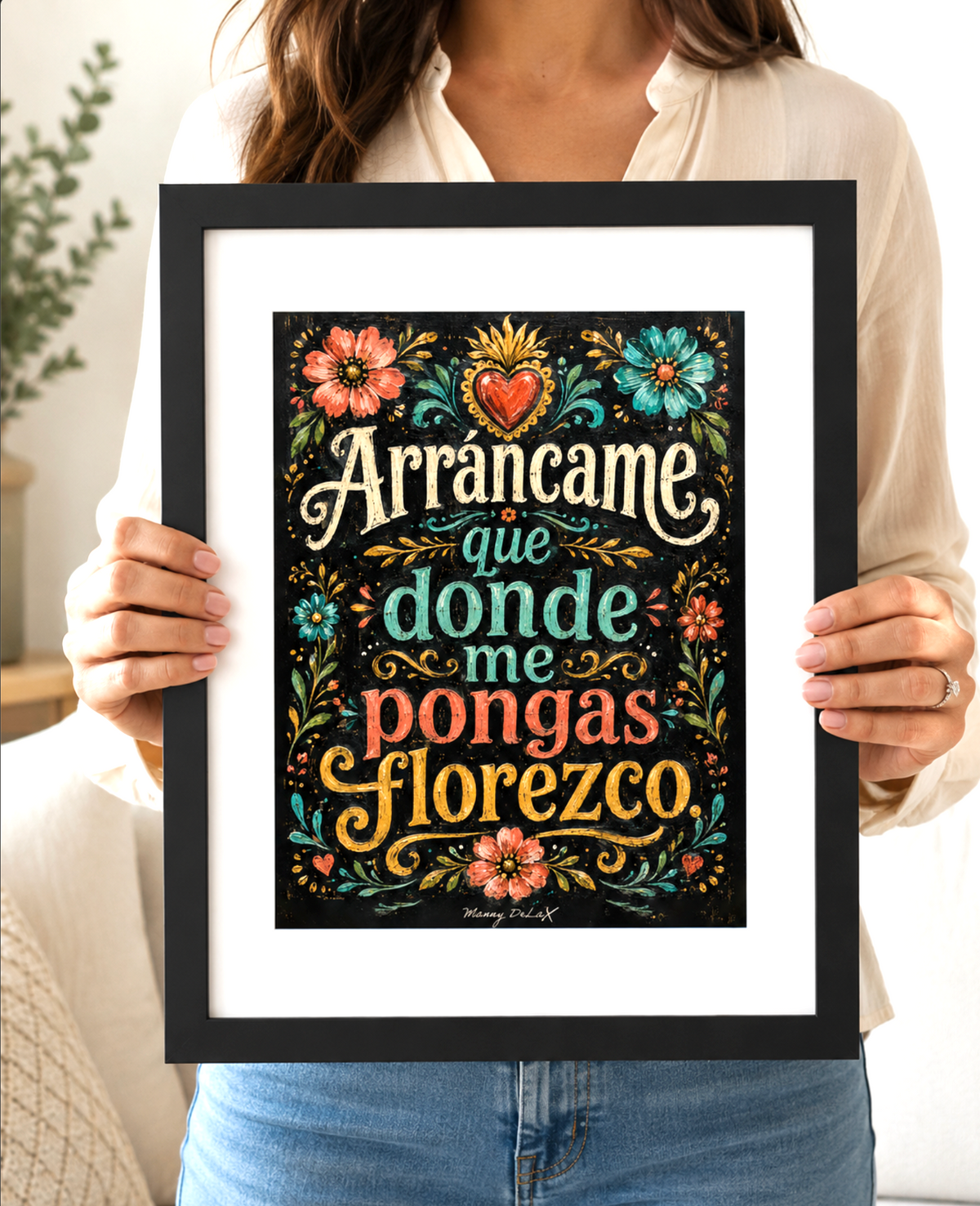 A person holding a black-framed art print featuring the Spanish phrase 'Arrancame que donde me pongas florezco' in decorative multicolor lettering with floral and heart motifs on a black background.
