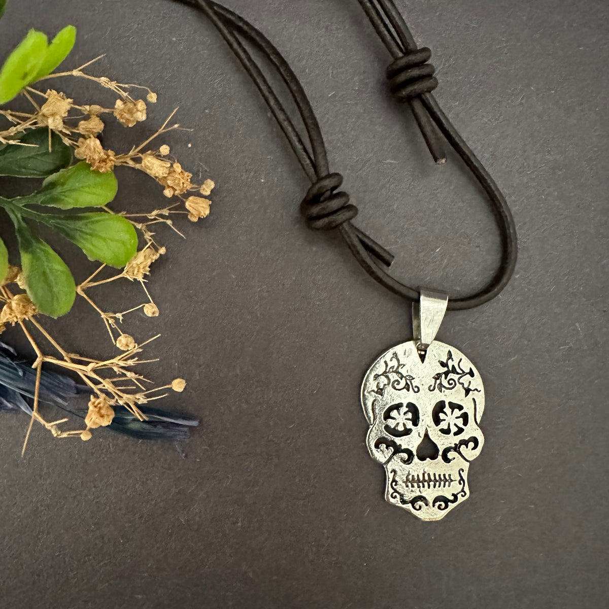 Sugar skull necklace arranged vertically on gray, with the silver-tone pendant below two sliding knots in the brown leather cord.