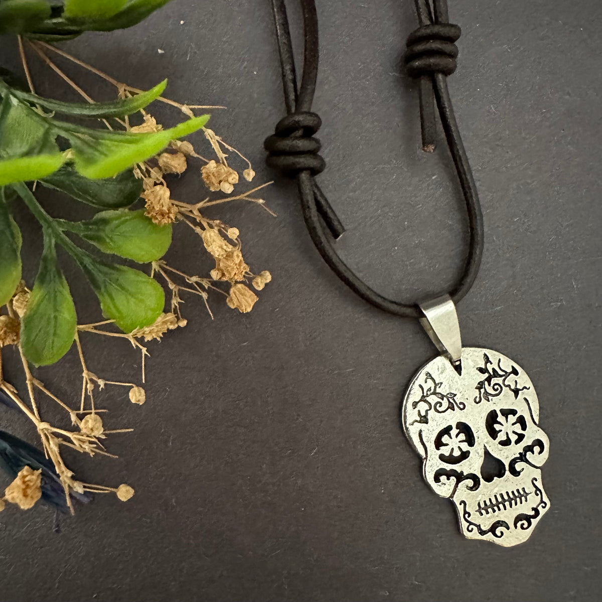 Front view of the silver-tone skull pendant below a brown leather cord, showing floral eye openings, scrollwork, and teeth beside greenery.
