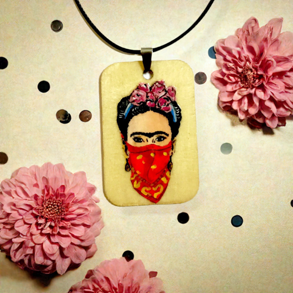 Frida-inspired necklace with a rectangular pendant depicting a woman wearing pink flowers and a red bandana, suspended from a black cord.