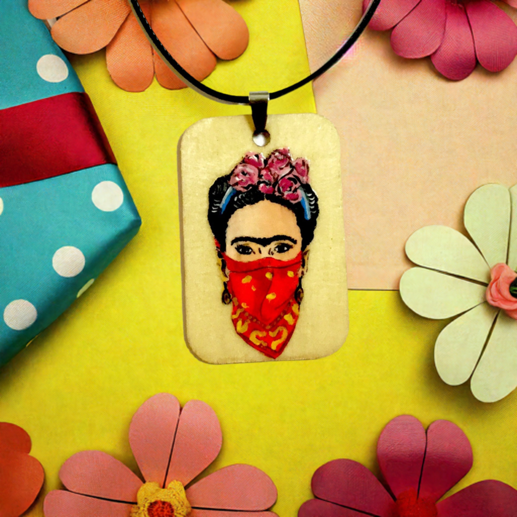 Frida-inspired necklace with a rectangular pendant depicting a woman wearing pink flowers and a red bandana, suspended from a black cord.