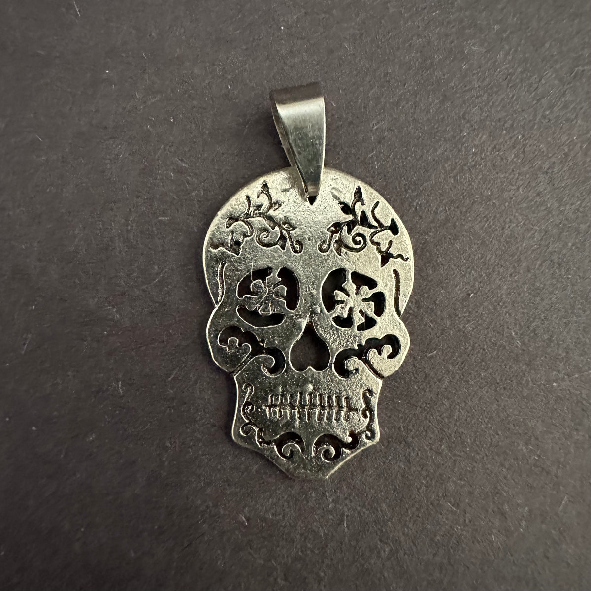 Silver-tone sugar skull pendant alone on a gray surface, with flower-shaped eye cutouts, dark scrollwork, defined teeth, and a tapered bail.

