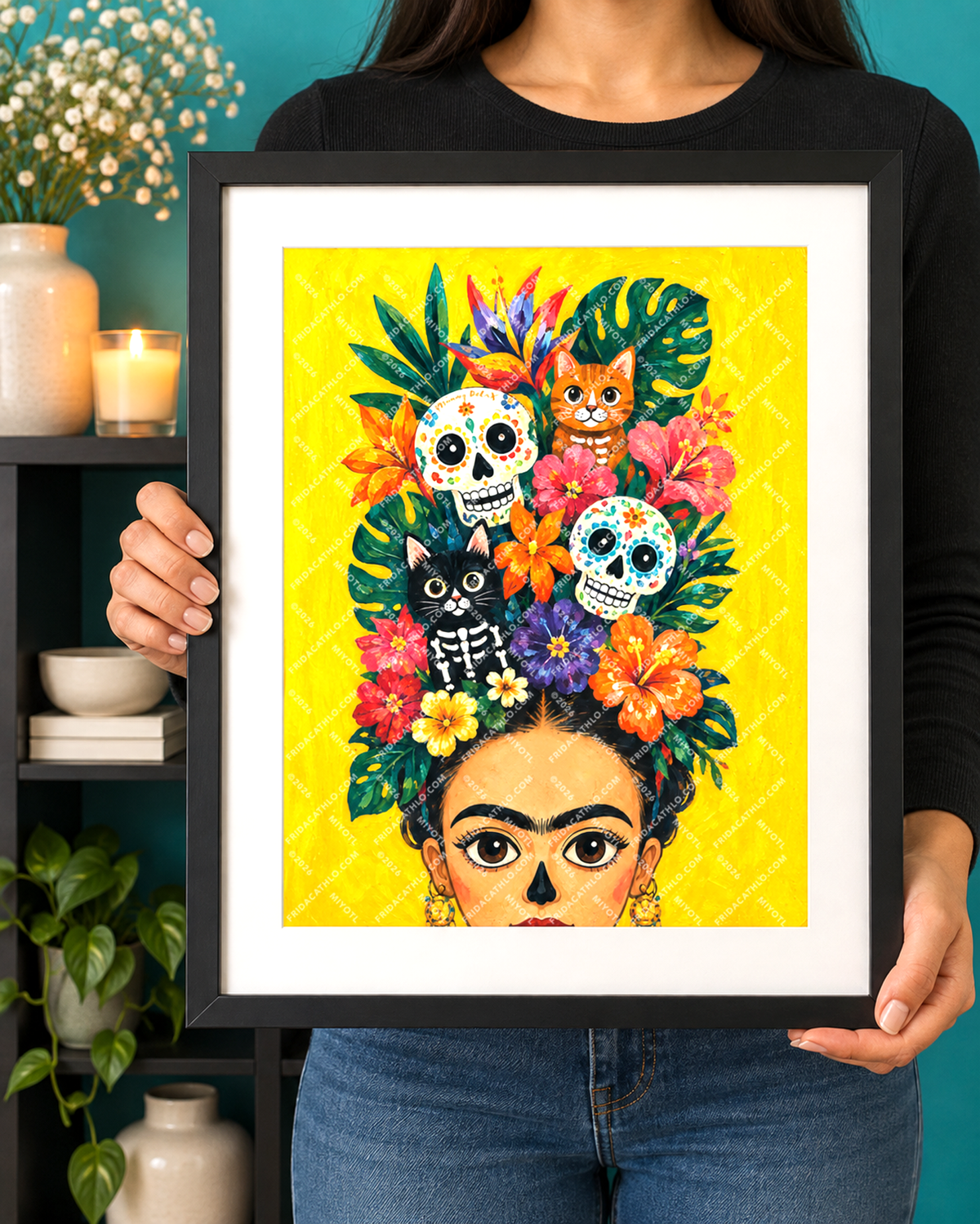 A person holds a framed art print featuring a colorful Day of the Dead style portrait of Frida Kahlo with flowers, skulls, and cats on a bright yellow background. The person is wearing a black long-sleeve top and blue jeans, standing in front of a teal wall with a shelf holding candles, flowers, and a plant.