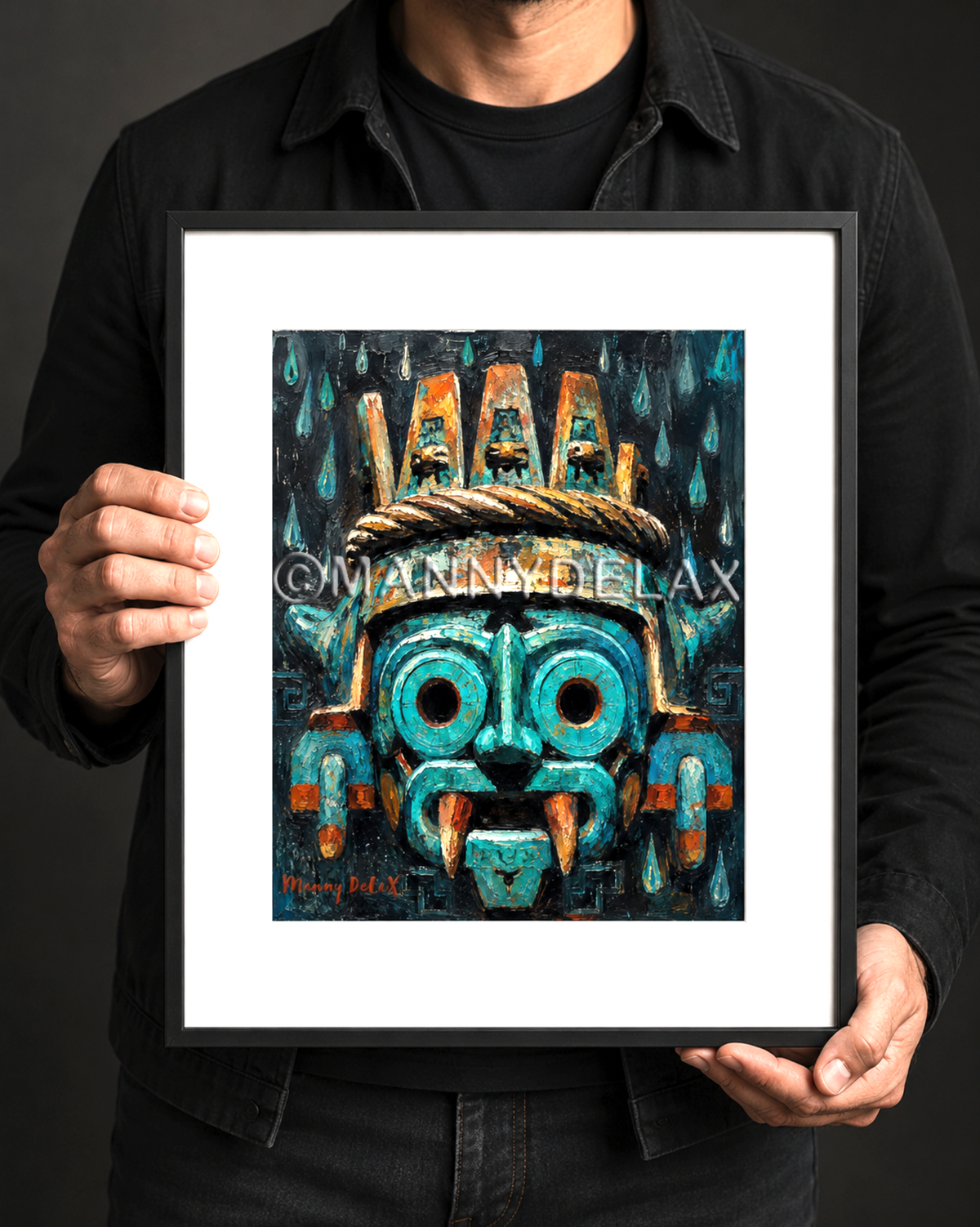 A person in a black shirt holds a framed art print featuring a stylized turquoise and orange Aztec-inspired face against a dark background.