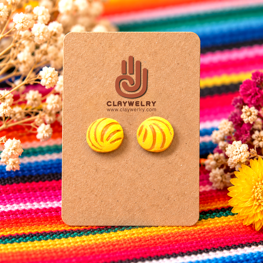 Yellow concha polymer clay stud earrings on a kraft display card against colorful striped fabric.
