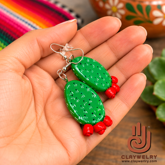 Green nopal cactus clay earrings with red flowers resting in an open palm.
