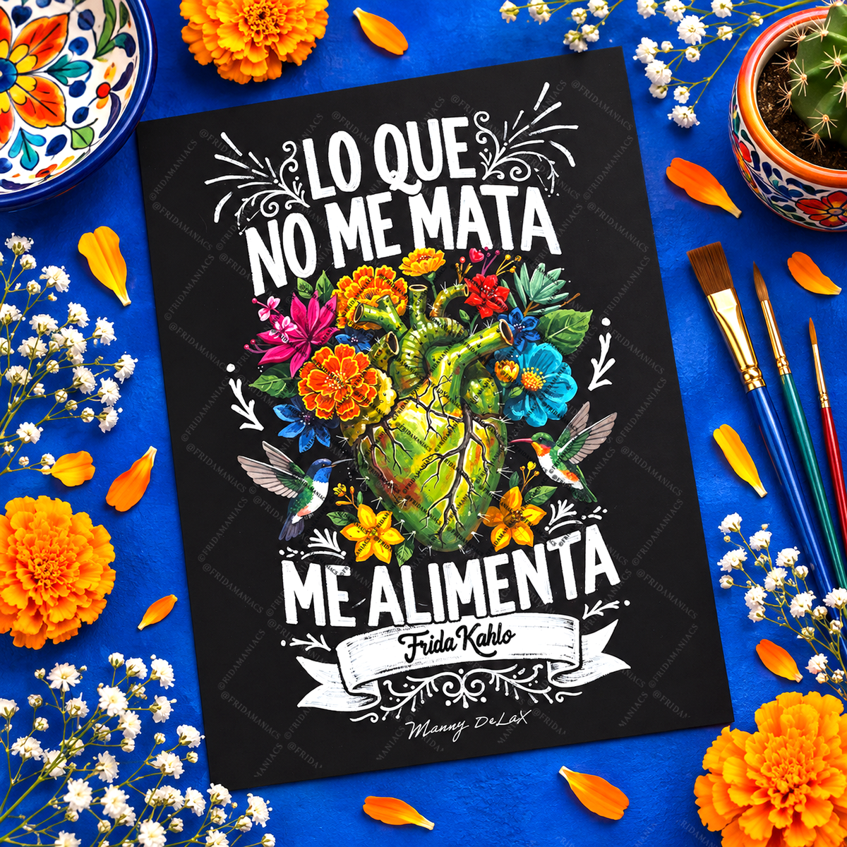 A black art print featuring a colorful illustration of a cracked green heart surrounded by flowers, hummingbirds, and marigolds, with the Spanish text 'LO QUE NO ME MATA ME ALIMENTA' and 'Frida Kahlo'. The print is displayed in a styled flat-lay on a bright blue background decorated with flowers, paintbrushes, and a small cactus.