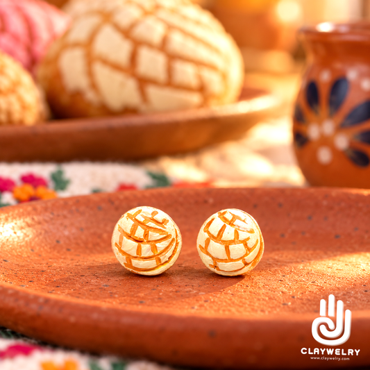 White vanilla concha clay stud earrings held on fingertips, showing metal posts and clear backs, with sweet bread and coffee behind.
