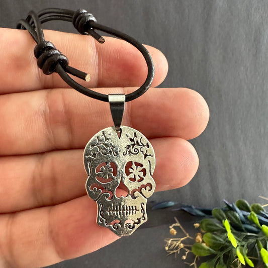 A man's necklace with a rustic silver sugar skull pendant and brown leather. Silver-tone sugar skull pendant resting against fingers, with flower-shaped eye cutouts and brown leather cord with sliding knots.
