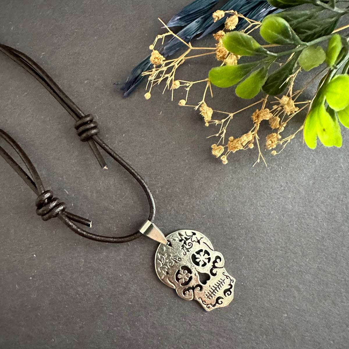 Brown leather skull necklace angled across a gray surface, with the silver-tone pendant at lower right and foliage above.
