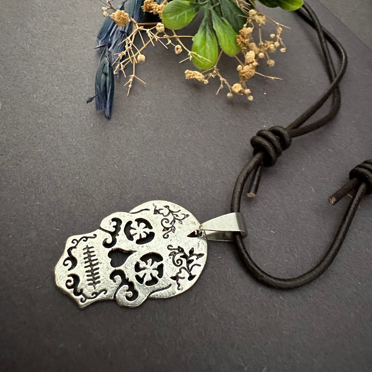 Rustic silver-tone sugar skull necklace lying diagonally on a gray surface, with brown leather sliding knots beside decorative foliage.
cords.