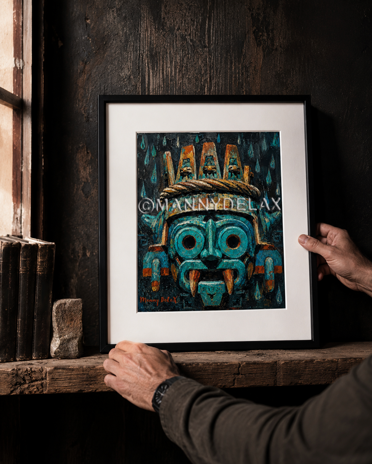 A framed art print featuring a stylized turquoise and orange Aztec-style mask, held by hands on a rustic wooden shelf against a dark wood wall.