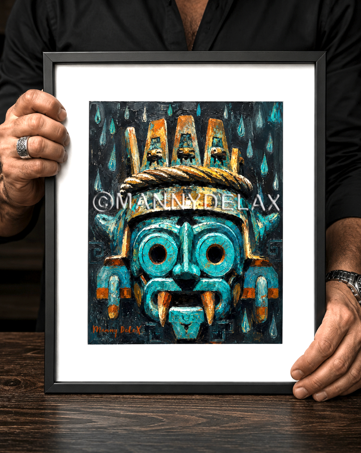 A framed art print featuring a stylized turquoise and gold Aztec-inspired mask, held by a person wearing a black shirt over a dark wooden table.