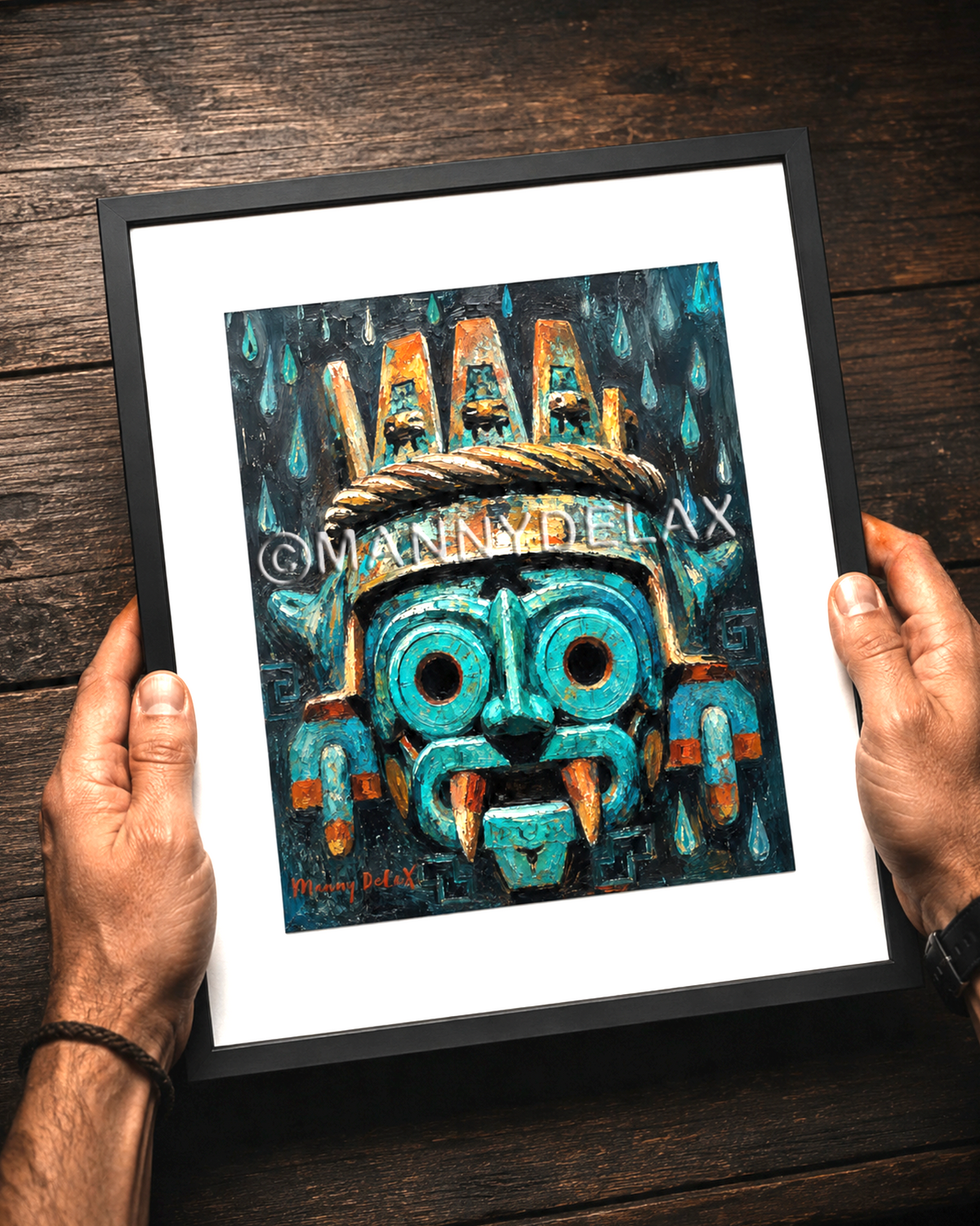 A framed art print featuring a stylized turquoise and orange Aztec mask under a rain motif, held by two hands over a dark wood background. The print has a white mat and a black frame.