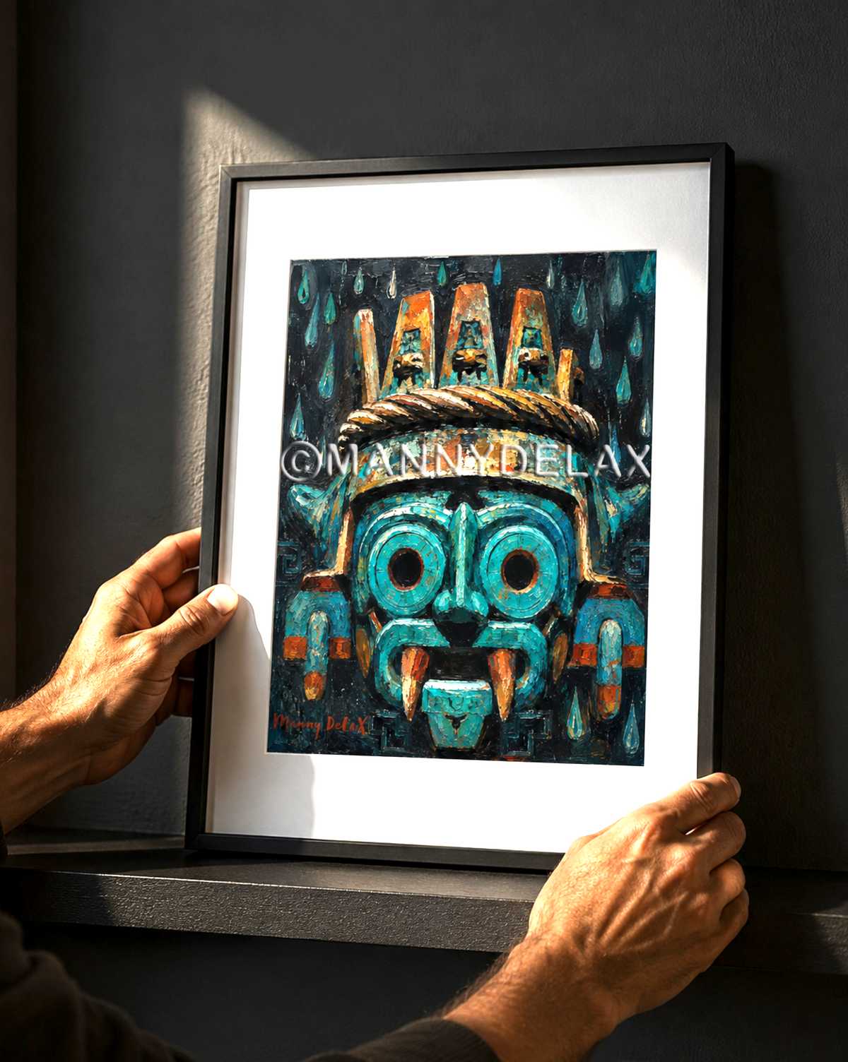 A framed art print featuring a stylized turquoise and orange Aztec-style mask, held by two hands against a dark gray wall.