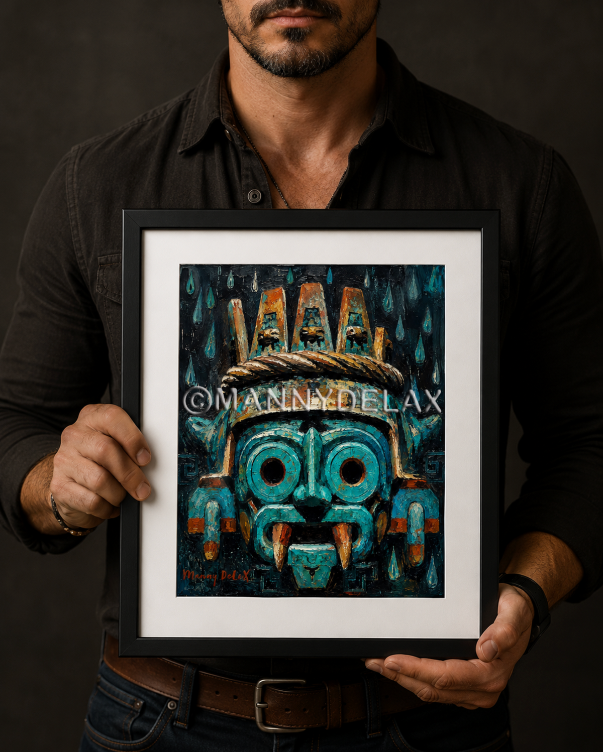 A man in a black shirt holds a framed art print featuring a stylized turquoise and black Aztec-style mask with gold details. The print has a white mat and a black frame, and the artist's name 'Manna Delax' is printed across the center.
