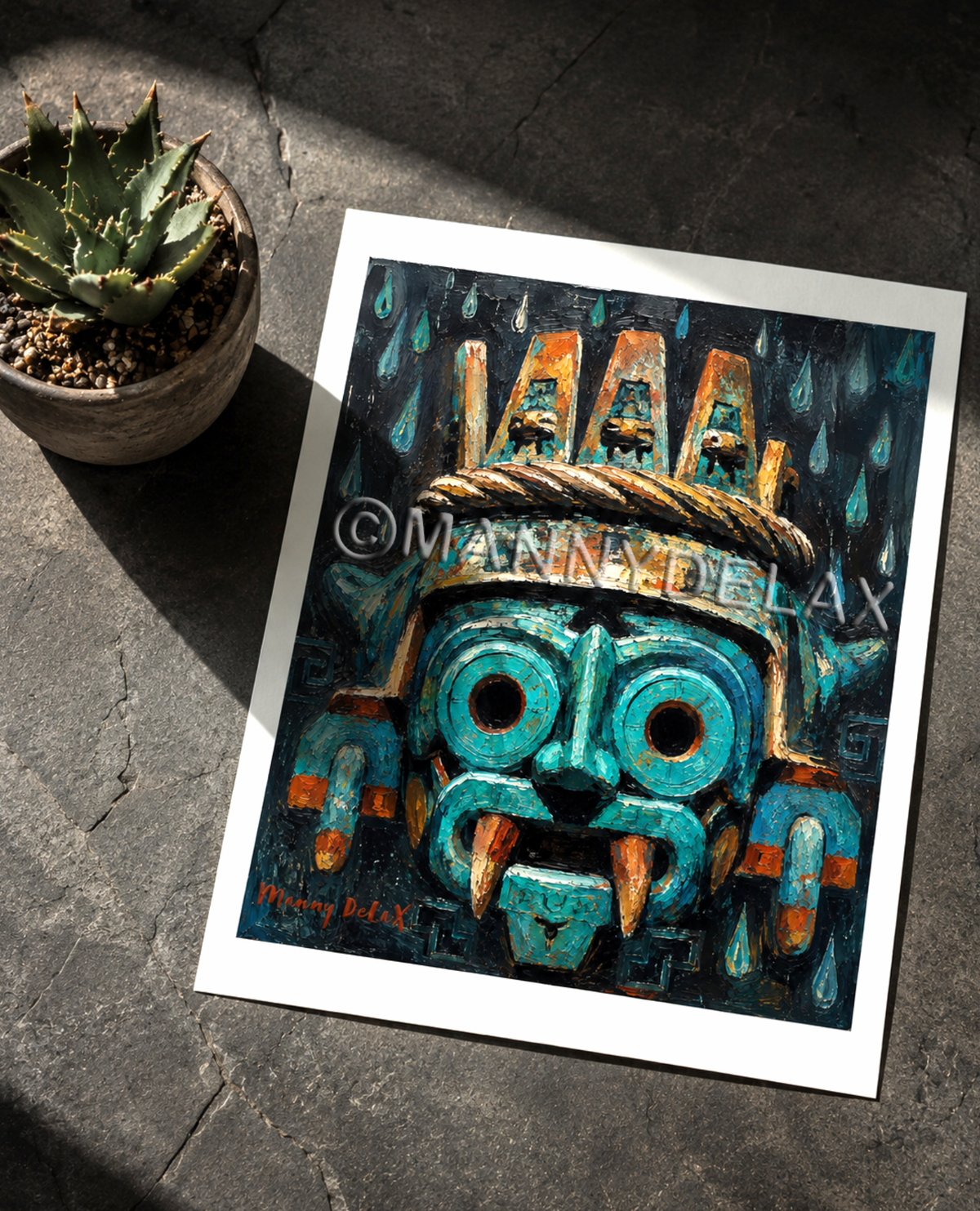 A square art print featuring a stylized turquoise and black Aztec-style mask with orange accents, photographed on a dark stone surface next to a small potted succulent. The print has a white border and a visible watermark reading '@MANNYDELAX'.