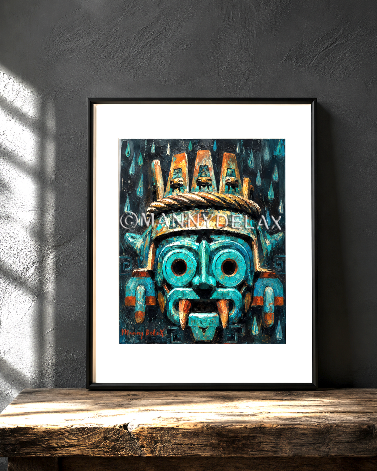 A framed art print featuring a stylized turquoise and black Aztec mask with gold accents, displayed on a rustic wooden table against a dark gray textured wall with soft window shadows.