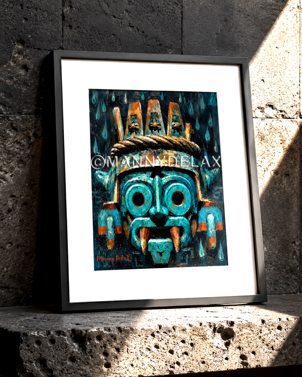 A framed art print featuring a stylized turquoise and orange Aztec-style mask [TLALOC] on a dark background, leaning against a textured concrete wall on a stone surface.