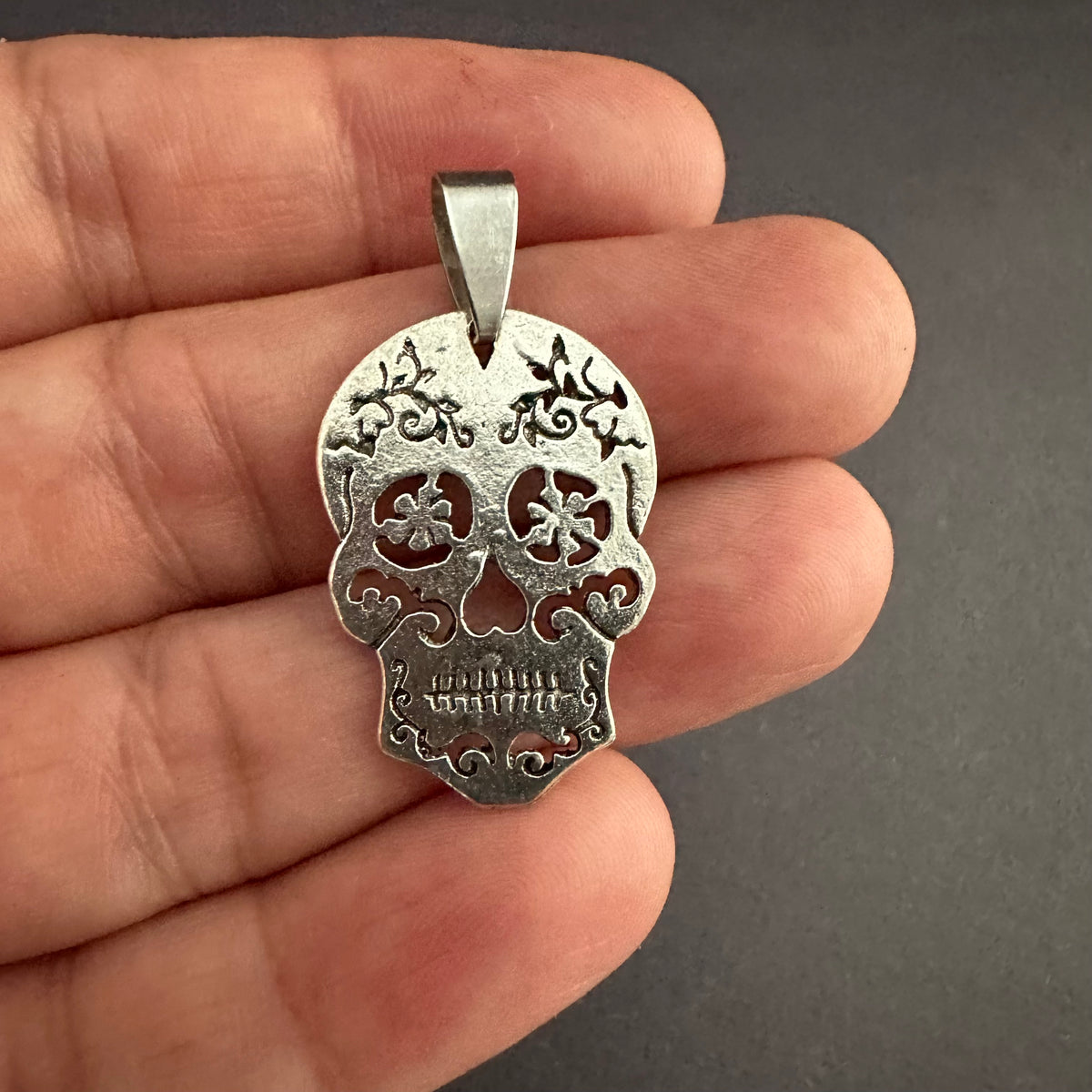 Front view of the silver-tone skull pendant against an open hand, showing its cutout eyes and nose, engraved details, and textured finish.