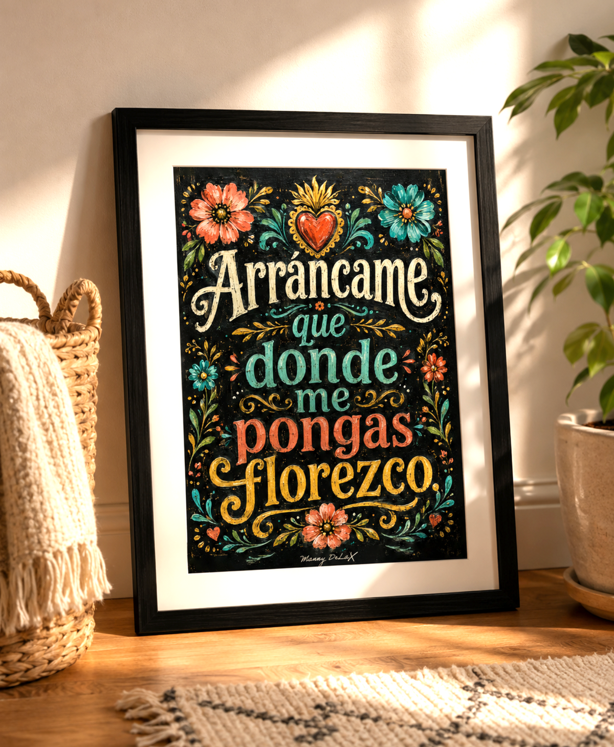 A framed decorative art print leaning against a wall on a wooden floor, with a woven basket and throw on the left and a potted plant on the right. The print features colorful floral illustrations and the Spanish text 'Arrancame que donde me pongas floresco' in stylized lettering.