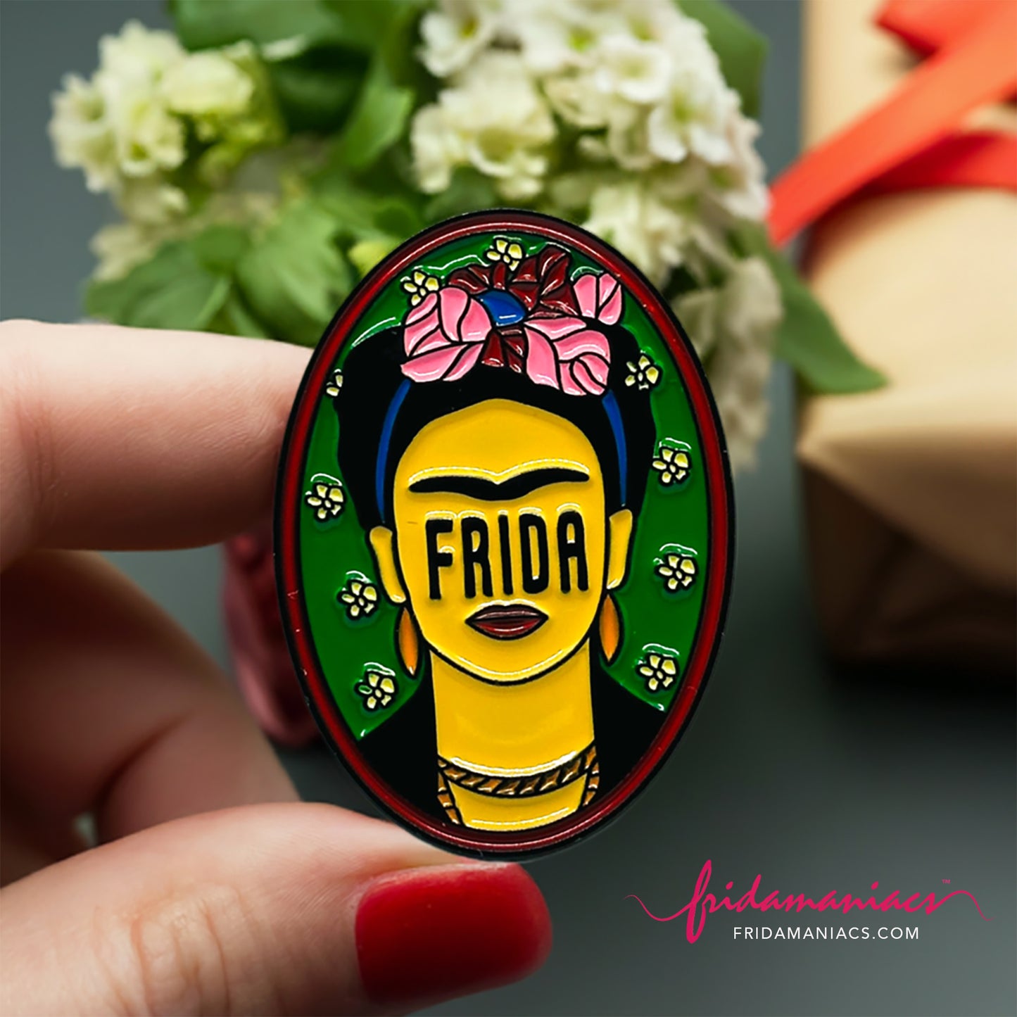 Frida Kahlo oval enamel pin back button with green enamel as background, wine tone frame, and FRIDA spelling instead of eyes below iconic artist eyebrows with pink flowers. Fridamaniacs. Mexican jewelry.