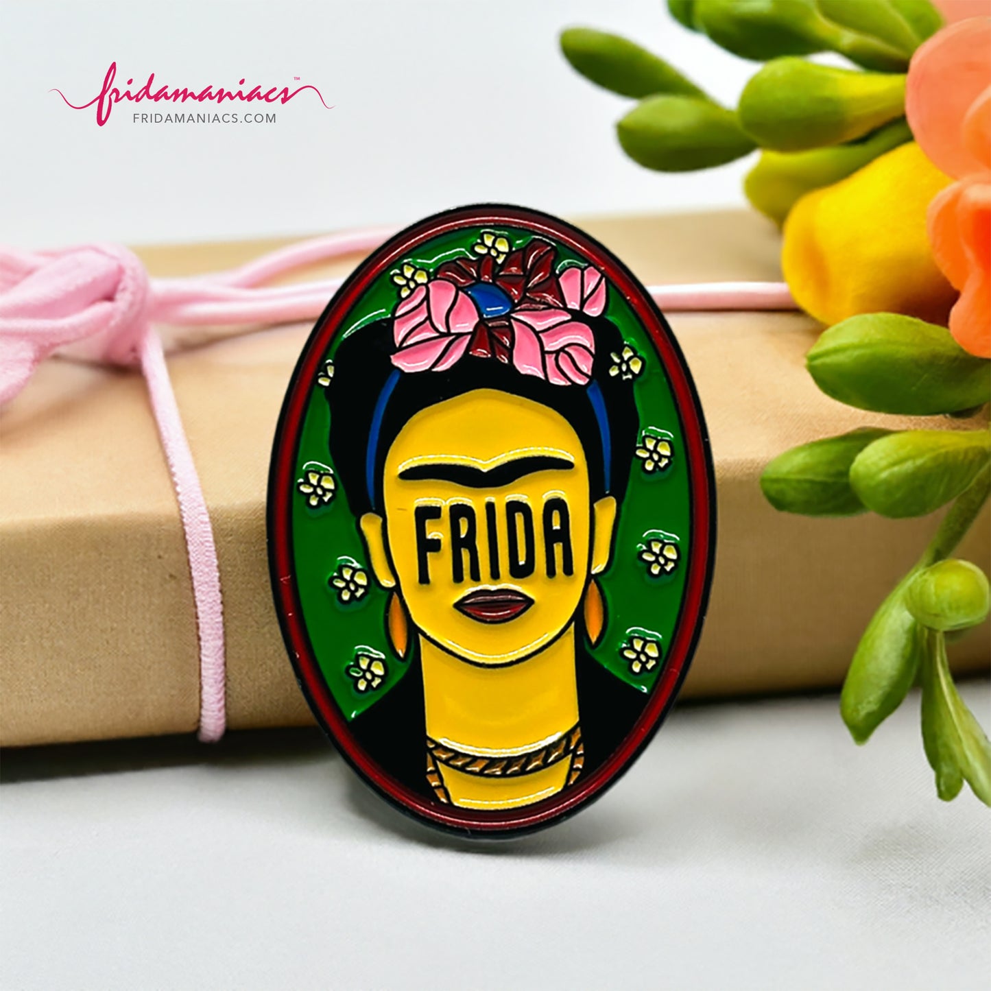 Frida Kahlo oval enamel pin back button with green enamel as background, wine tone frame, and FRIDA spelling instead of eyes below iconic artist eyebrows with pink flowers. Fridamaniacs. Mexican jewelry.