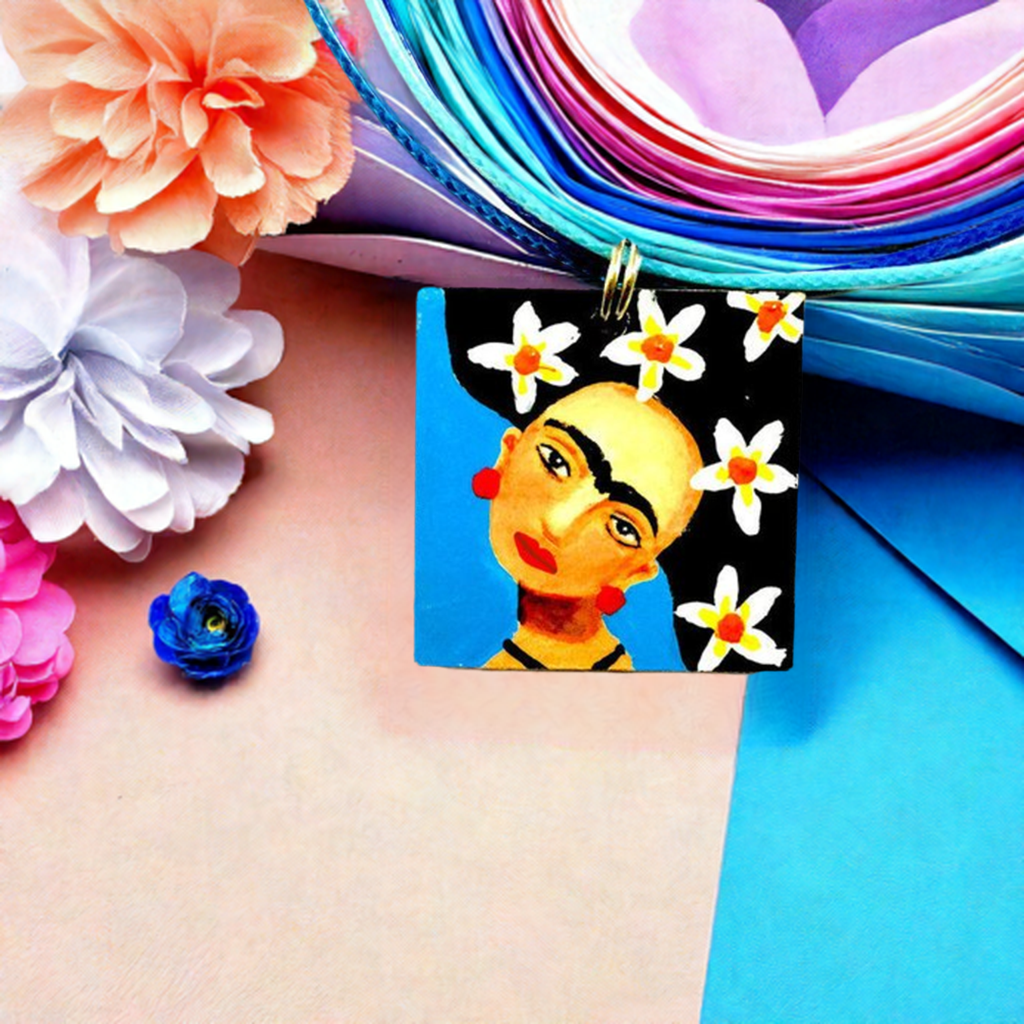 Hand painted Frida Kahlo inspired pendant with blue and white-yellow-orange wilds flowers on hair. Mexican artist portrait art to wear for women and girls. Unique gift idea for fridamaniacs, fridalovers, and frida fans.