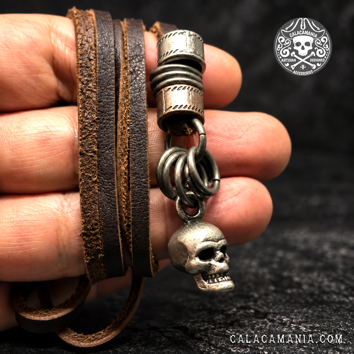 Skull Bonded leather necklace held against a black background: Brown bonded leather skull necklace held in a hand, showing textured cords and antiqued metal rings.

