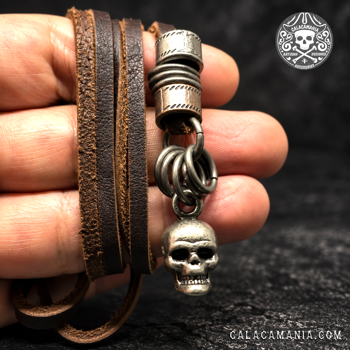 necklace held against a black background:
Brown bonded leather skull necklace held in a hand, showing textured cords and antiqued metal rings.
