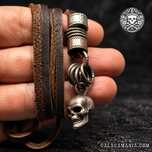 Necklace held against a black background: Brown bonded leather skull necklace held in a hand, showing textured leather cords and antiqued metal rings.
